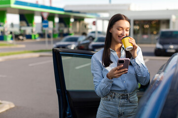 Woman car traveler standing near her car, drinking coffee from paper cup and using cellphone, stopped at the parking lot of gas station, taking break during road trip