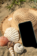 Smartphone on sand with shells and beach hat.