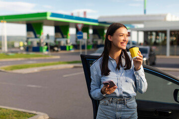 Happy lady holding cellphone and drinking coffee, standing near her car on the petrol station parking, having break during road trip, copy space