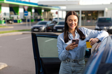 Young happy woman standing near her car, smiling and using cellphone, holding coffee cup, making...