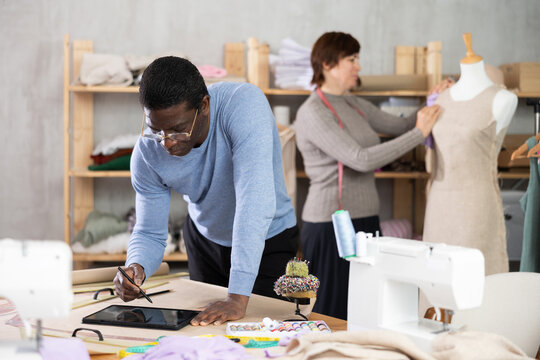 Focused experienced fashion designer working on digital sketch of new garment using tablet in studio while female colleague working on fitting dress on mannequin in background