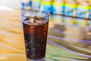 Glass of cola with ice and visible condensation on surface standing on colorful reflective table with background blur. Aruba.