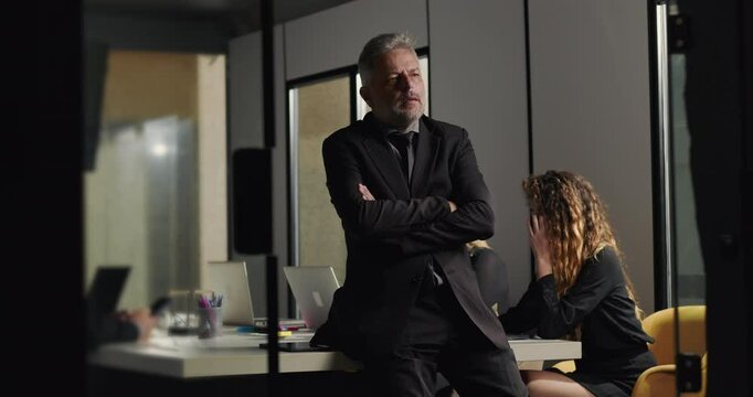 Senior businessman standing with thoughtful pose in office boardroom while coworkers concentrate on laptops during company meeting