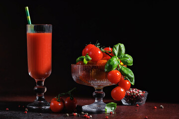 Healthy cherry tomatoes with basil leaves in a glass dessert cup next to a tall drink of tomato juice on a red rustic surface