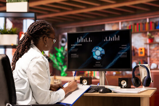 Remote worker at home watching business conference on PC monitor, writing down info. Freelancer in apartment office looking at presentation graphs on computer, taking notes