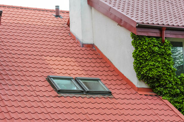 Close-up of red clay roof tiles and skylight window with green ivy growing on white residential...