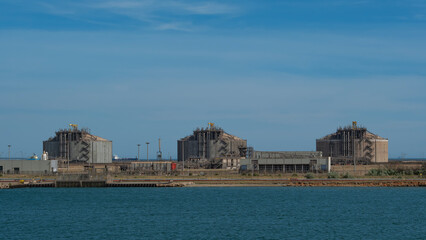Three large industrial tanks with complex structures at a port, situated by the water under a vast, clear blue sky
