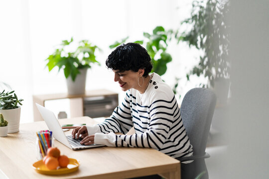 A woman is working on her laptop at a desk in a bright, plant-filled room. She's smiling, likely enjoying her work or connecting with someone online.