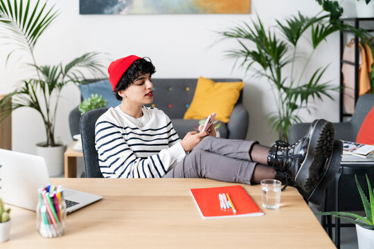 A young woman in a red beanie takes a break from work. She's checking her phone, feet up on the desk, surrounded by plants in her home office.