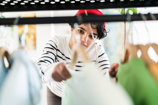 A woman in a striped shirt and red beret is browsing clothes on a rack. She's carefully looking through the items, perhaps searching for a specific style or color.