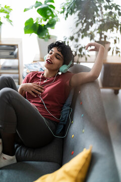 A woman is relaxing on her couch, listening to music with headphones. She's enjoying the moment and feeling the rhythm, maybe after a long day.