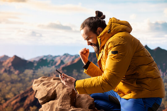 Male hiker reading some good news and feeling joyful with his phone, being distracted by the strong signal and internet connection. Modern explorer cheering with excitement on mountain peak.