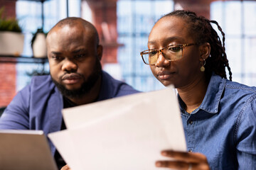 Young remote workers examining calendar appointments on their documents, checking papers and laptop information to solve important tasks on schedule. Couple collaborating at home.