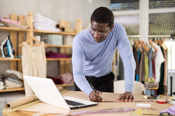 Male tailor draws sketch of future clothes on paper while checking data from laptop in a sewing workshop