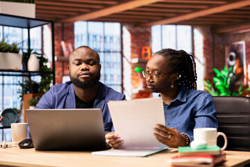 Black remote employees organizing calendar appointments using laptop, checking their papers and synchronizing important tasks on schedule. Man and woman couple review files at home.