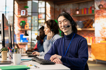 Portrait of happy call center consultant handling customer inquiries, complaints and support requests in office. Cheerful Asian man doing customer service job assisting clients with issues related to