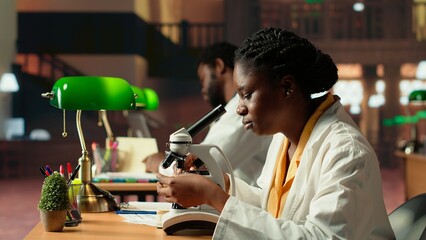 African american future researcher prepares for exam in a quiet study space, examining biological cells on a microscope. Woman student takes personal notes, career in medical science. Camera B.