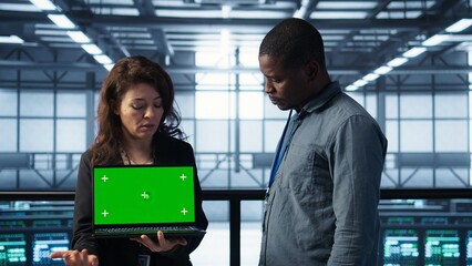 Data center IT team automating routine maintenance tasks with green screen laptop, executing diagnostic scripts. Server room coworkers using mockup notebook, adjusting power settings, camera B