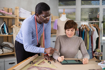 Two interested fashion designers, man and woman, collaborating in sewing studio, using tablet to create and refine new garment design at pattern table, discussing digital sketches or client references