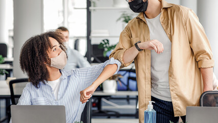 Obraz premium Hello colleague and new normal. Arabian guy and african american woman in protective masks greet with elbows at workplace in interior of modern office during coronavirus epidemic, copy space