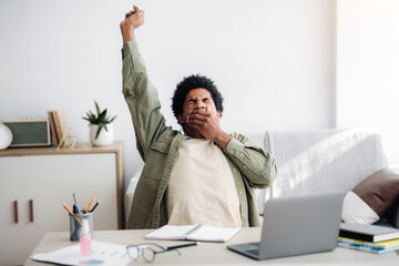 Tired black student yawning and stretching during his remote studies from home. African American youth exhausted from getting ready for test or writing coursework, feeling sleepy in front of laptop
