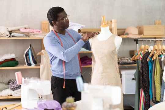 African American man tailor connects parts of dress on mannequin. Female craftsman attaches front part of dress to back with pins.