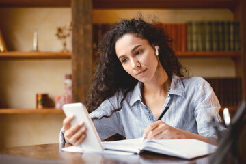 Remote Learning. Portrait of young woman in wireless earbuds sitting at desk, using smart phone and...