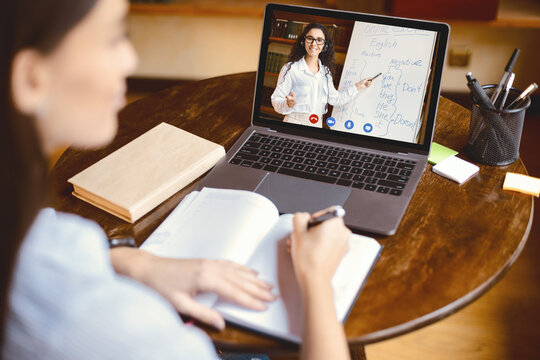 E-learning Concept. Over the shoulder back view of female student sitting at table, writing in notebook, taking notes, using laptop, having online class with English teacher. Distant studying