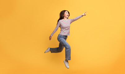 Obraz premium Asian young woman is joyfully leaping in the air while pointing at copy space. She wears a striped long-sleeve shirt and jeans, radiating happiness and energy in a vibrant yellow space.