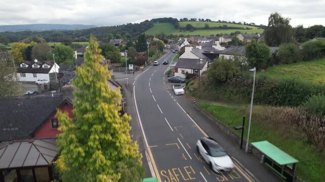 traffic on road passing by houses in village. Descending shot of moving vehicles and revealing bus stop. Wales, UK