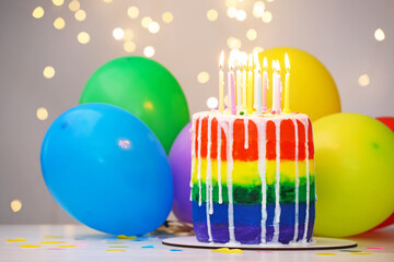 Delicious rainbow birthday cake with burning candles, colorful balloons and confetti on white table against grey background with blurred lights, closeup