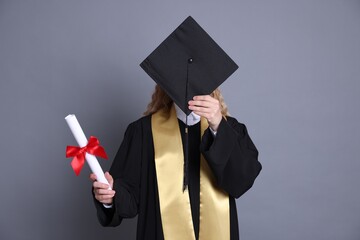 Student with diploma hiding behind mortarboard after graduation on grey background