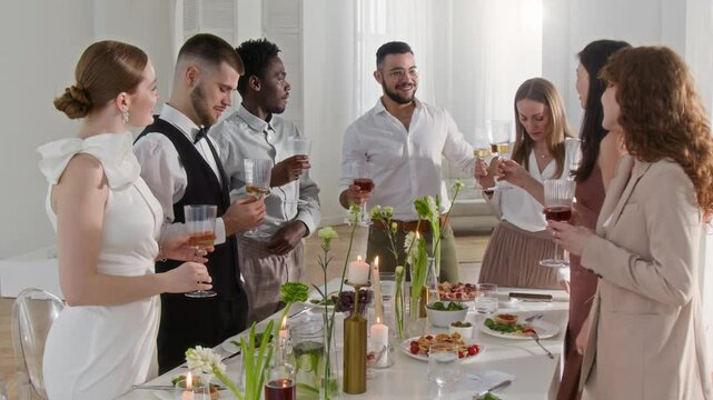Best man hugging his lover while standing with glass of wine and proposing toast at wedding. Guests and newlywed couple listening to his speech and enjoying moment