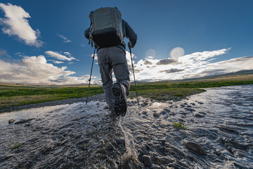 A photo of a man with a butt. A traveler walks on the water in trekking boots and with a backpack on his back. There is a green meadow and blue sky on the background. Waterproof boots.