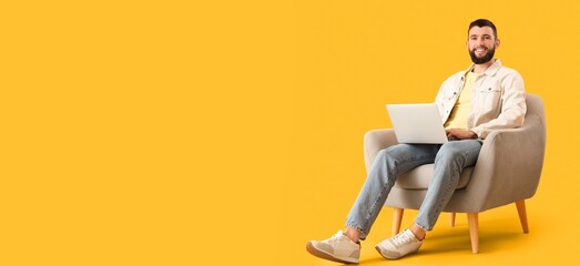 Handsome young man with laptop sitting in armchair against yellow background with space for text