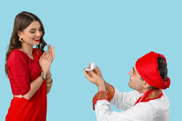 Young Indian man with engagement ring making proposal to his beloved girlfriend on blue background