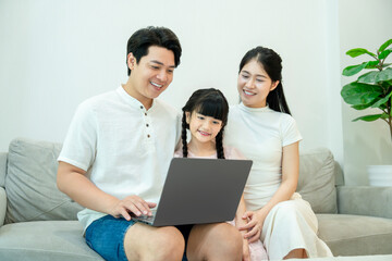 Fototapeta premium A relaxed Asian family sitting together on the couch, happily watching or shopping online using a laptop computer.