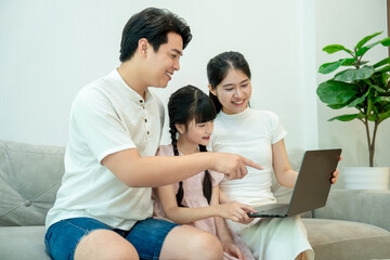 Fototapeta premium Attentive Asian parents are both pointing at a laptop screen, teaching their young daughter or showing her something interesting online.