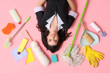 Happy young housemaid with different cleaning supplies lying on pink background