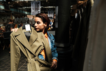 Young woman choosing trench coat in second hand clothing store