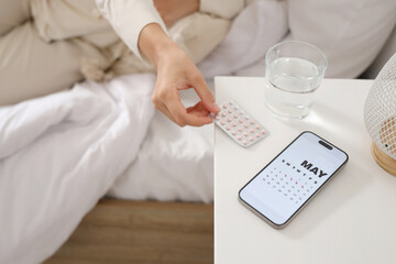 Young woman taking oral contraceptive pills and mobile phone with menstrual calendar on table in bedroom, closeup