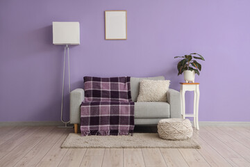 Sofa with stylish plaid, lamp and houseplant on stool near purple wall in room