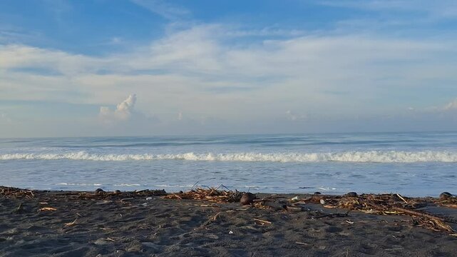 A serene tropical beach scene with soft waves crashing onto a black sand shoreline. The sky is bright and blue with scattered clouds, evoking a peaceful, natural atmosphere. Perfect for themes of rela