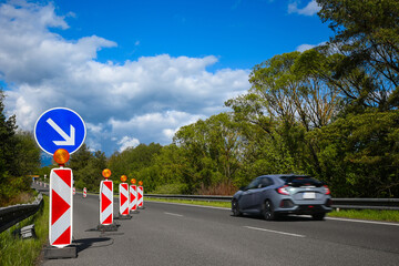 Highway lane closed for construction with diversion arrow sign, cones and flashing lights, car passing by on sunny day