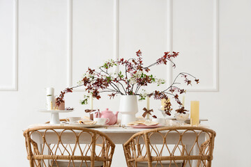 Dining table with blossoming branches and candles near light wall in room
