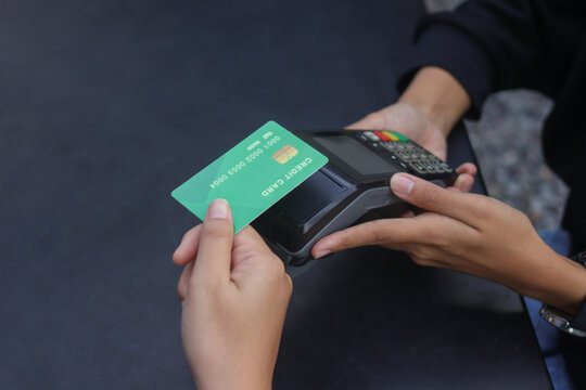 Credit card payment using a point of sale terminal. Close-up of hand swiping a green card while another person waits at a table. Modern cashless transaction concept.