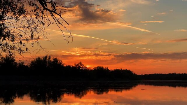 A colorful sunset in the picturesque sky over the Volga near Astrakhan. The sun disk enters the water. A beautiful summer or spring sunset on the Volga with trees reflected in the water. 4К