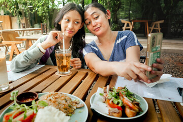 Young Asian mother and her teenage daughter smiling and taking selfie together using smartphone while having lunch at an outdoor restaurant. Close family relationship concept.
