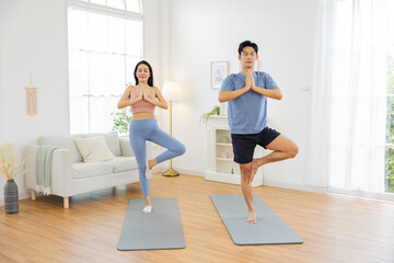 Obraz premium Asian man and woman practicing yoga together in bright living room, standing in tree pose on yoga mats, promoting mindfulness and balance in serene environment