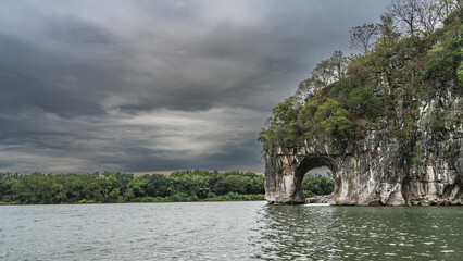 A picturesque rock with a rounded arched opening rises above the surface of the river. Green vegetation on the cliff slopes and on the shore. Dark clouds in the sky. China. Elephant Trunk Hill. Guilin